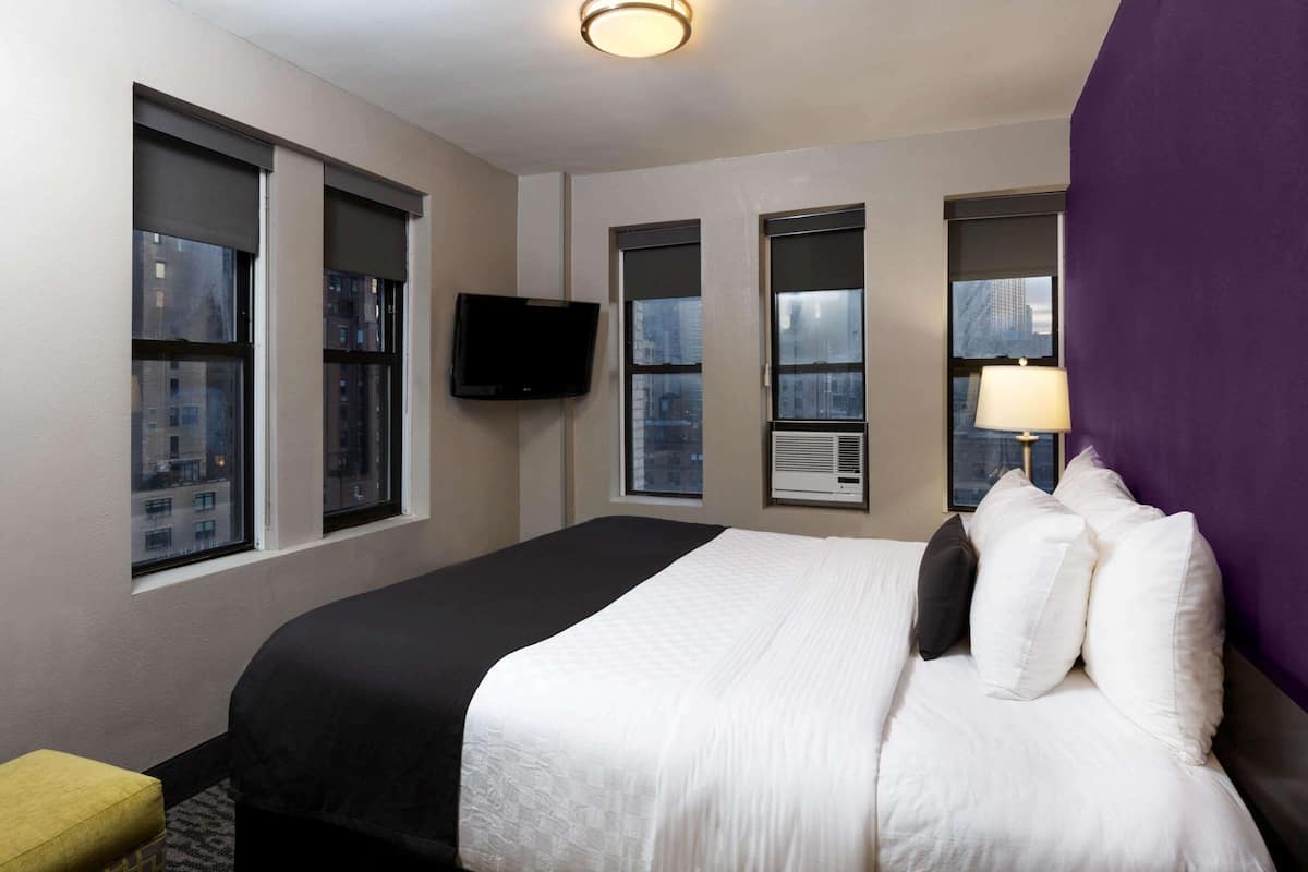 Une chambre d’hôtel avec un lit, un téléviseur fixé au mur et des fenêtres équipées de stores.