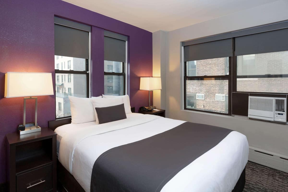 Une chambre d’hôtel avec un grand lit, deux tables de chevet avec des lampes, une fenêtre avec des stores et un appareil de climatisation.