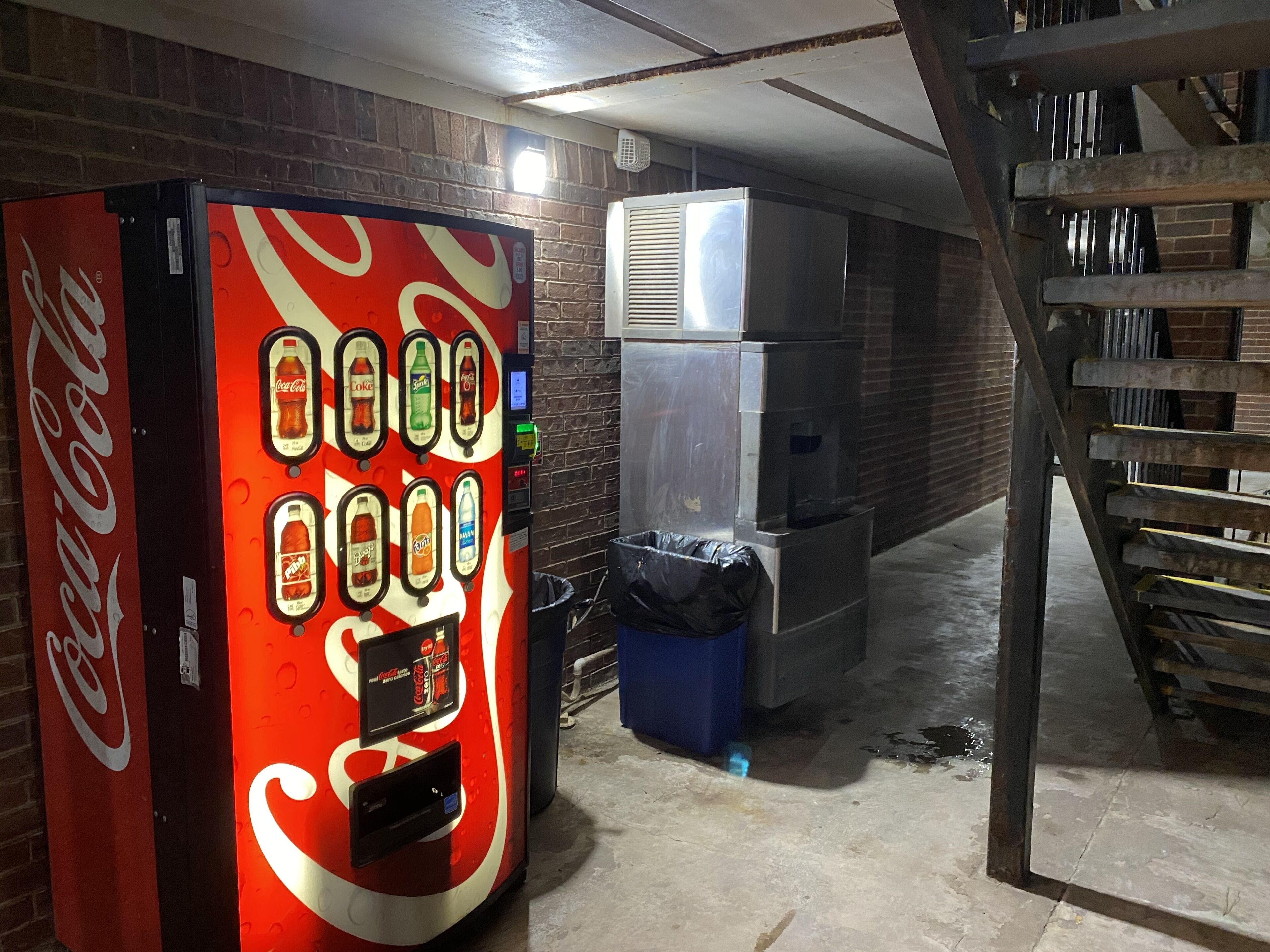 A vending machine with various drink options, a trash bin, and a staircase in a dimly lit area with brick walls.