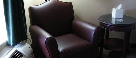 A hotel room with a leather armchair, a small round table with a tissue box, a lamp, and a painting on the wall.