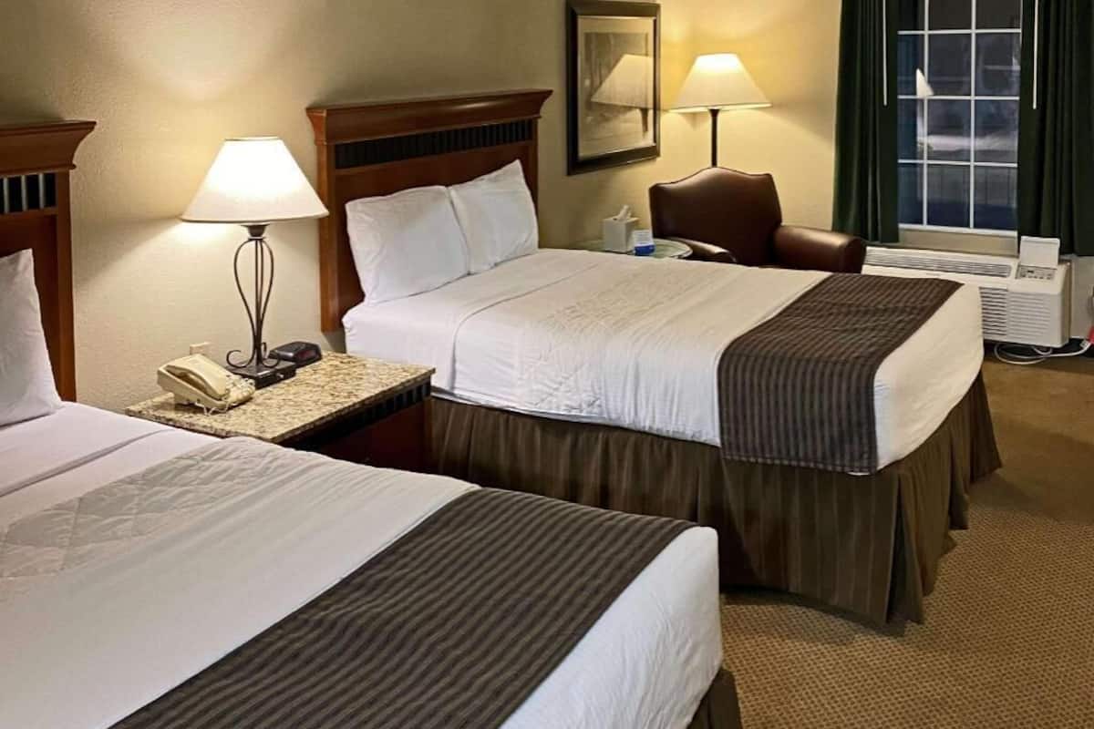 A hotel room with two beds, a nightstand with a lamp, a chair, and a window with curtains.