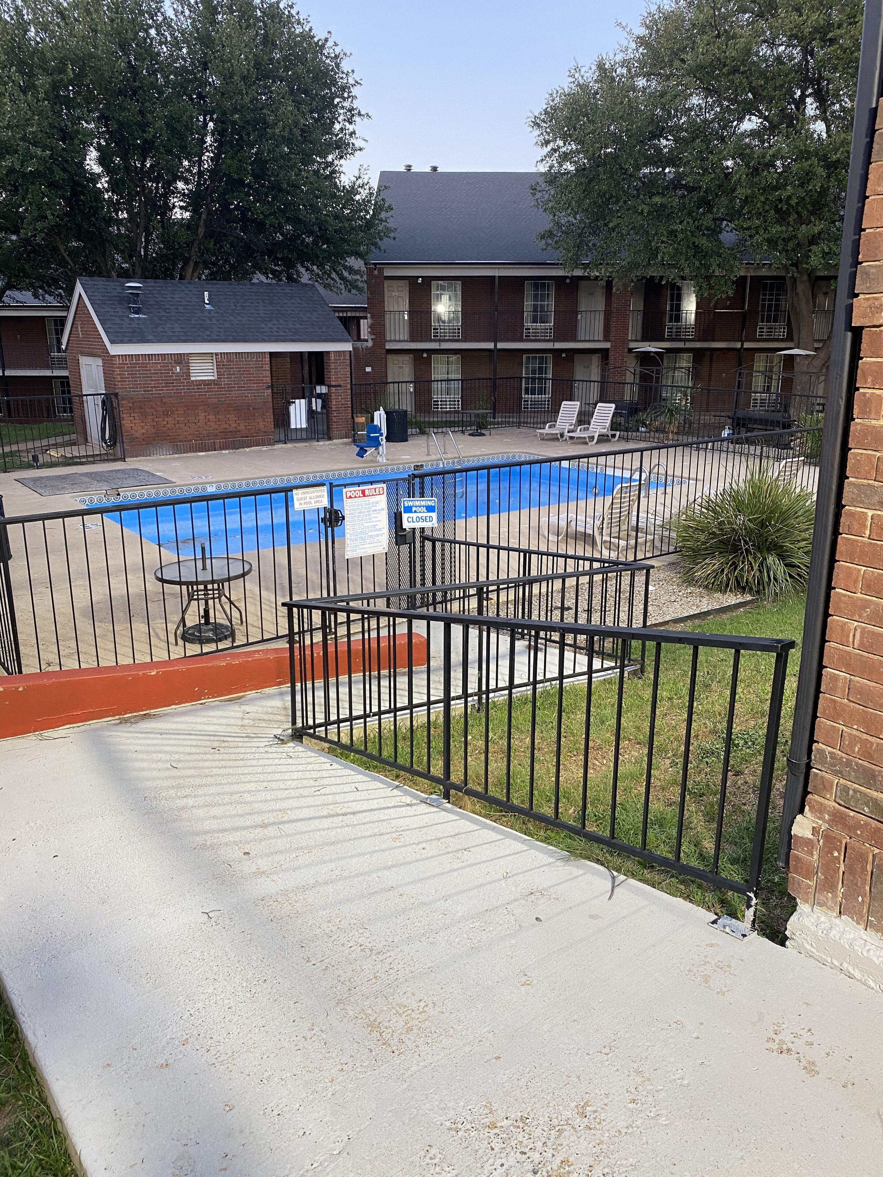 Une zone avec piscine, agrémentée d’un panneau indiquant que la piscine est fermée, d’une terrasse délimitée par une clôture et d’un bâtiment en briques en arrière-plan.