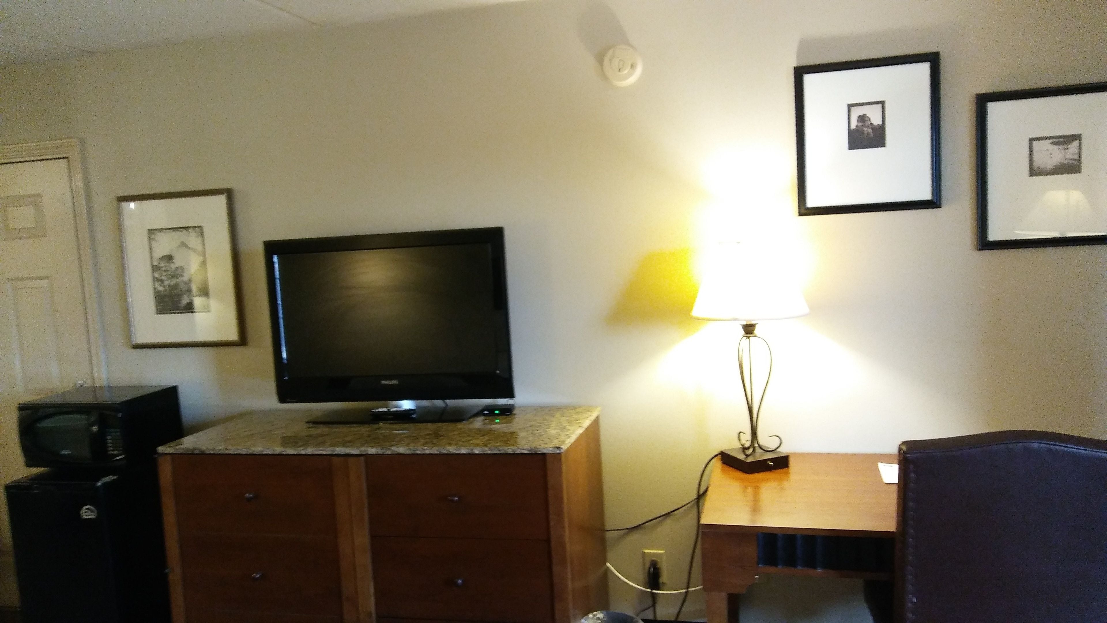 A hotel room with a flat-screen TV on a wooden cabinet, a desk with a lamp, and framed pictures on the wall.