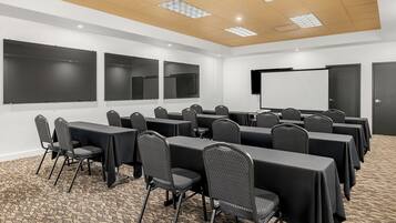 Un salón de conferencias con varias mesas y sillas, una pantalla de proyector y piso alfombrado.