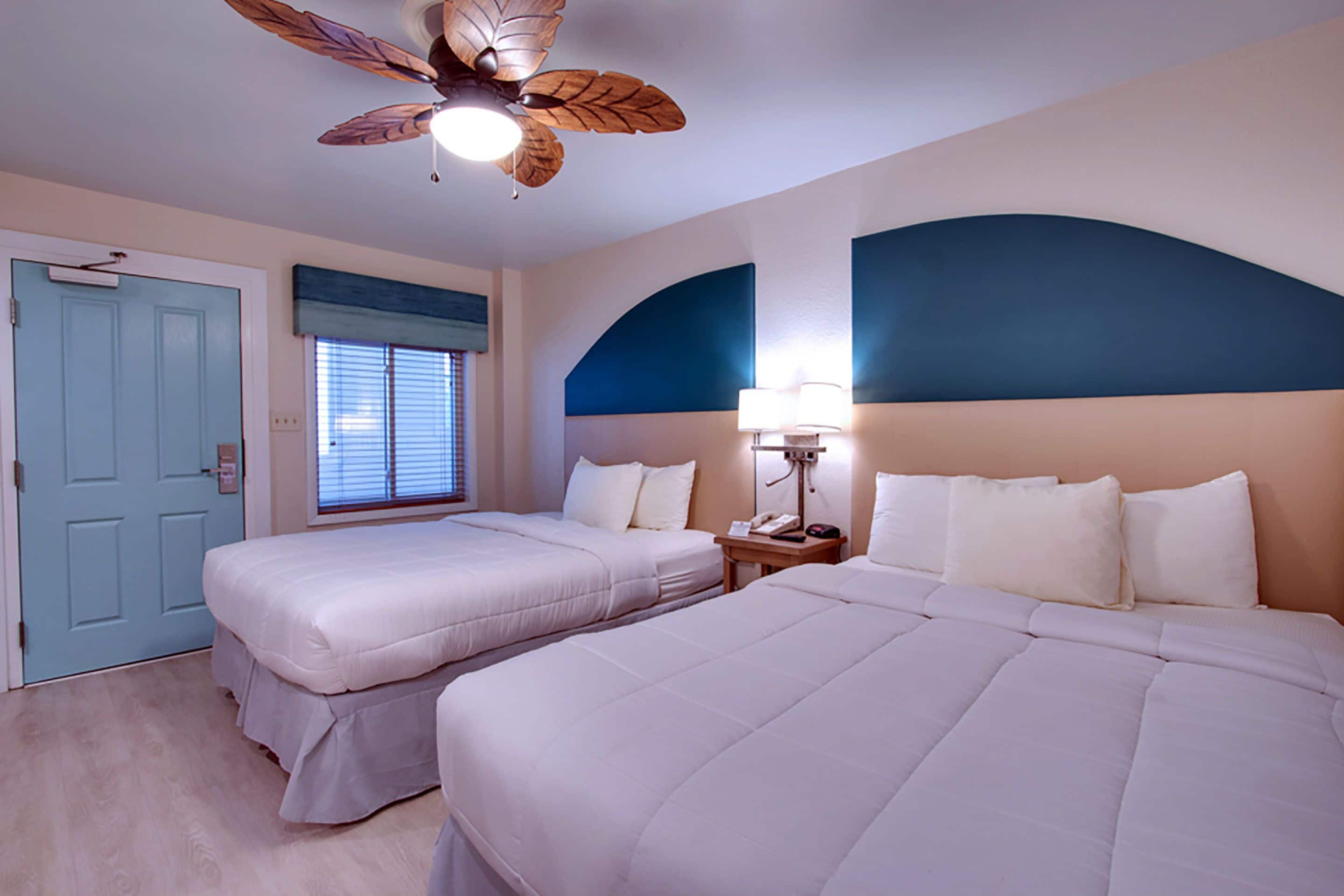 A hotel room with two beds, a ceiling fan, a nightstand with a lamp, and a window with blinds.
