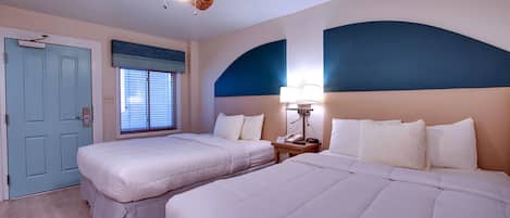 A hotel room with two beds, a ceiling fan, a nightstand with a lamp, and a window with blinds.