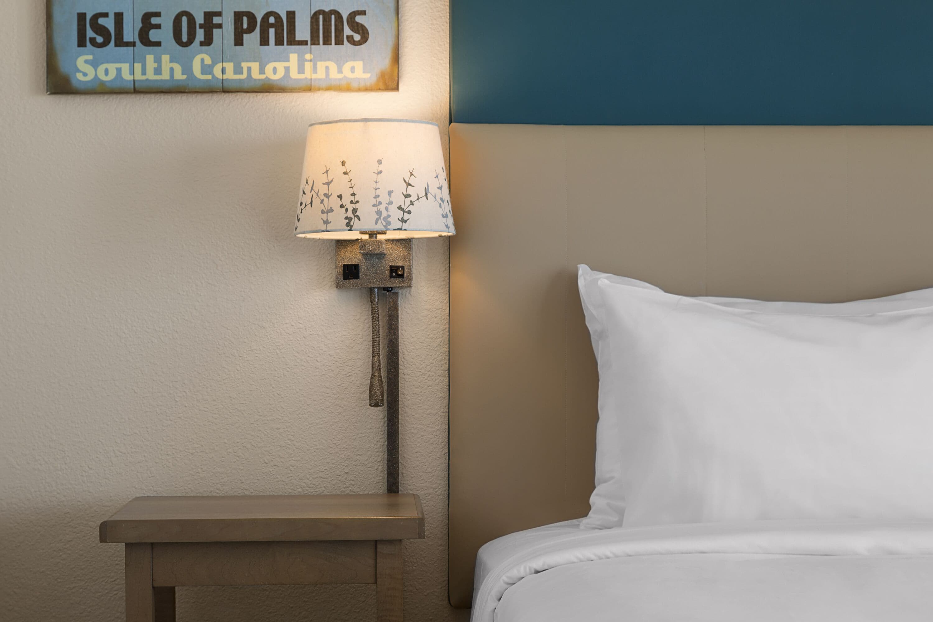 Ein Hotelzimmer mit einem Bett, einem Nachttisch, einer Lampe und einem Schild mit der Aufschrift 'ISLE OF PALMS South Carolina'.
