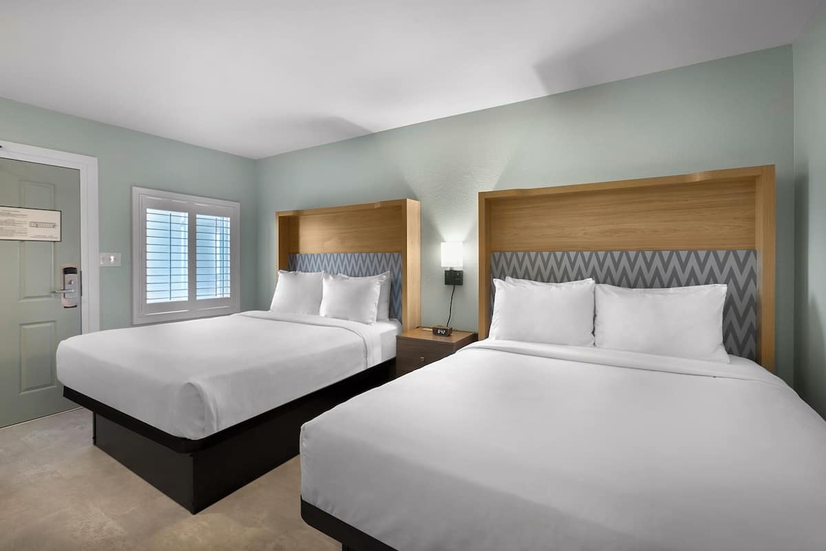 A hotel room with two beds, a wooden headboard, a nightstand, a lamp, and a window with blinds.