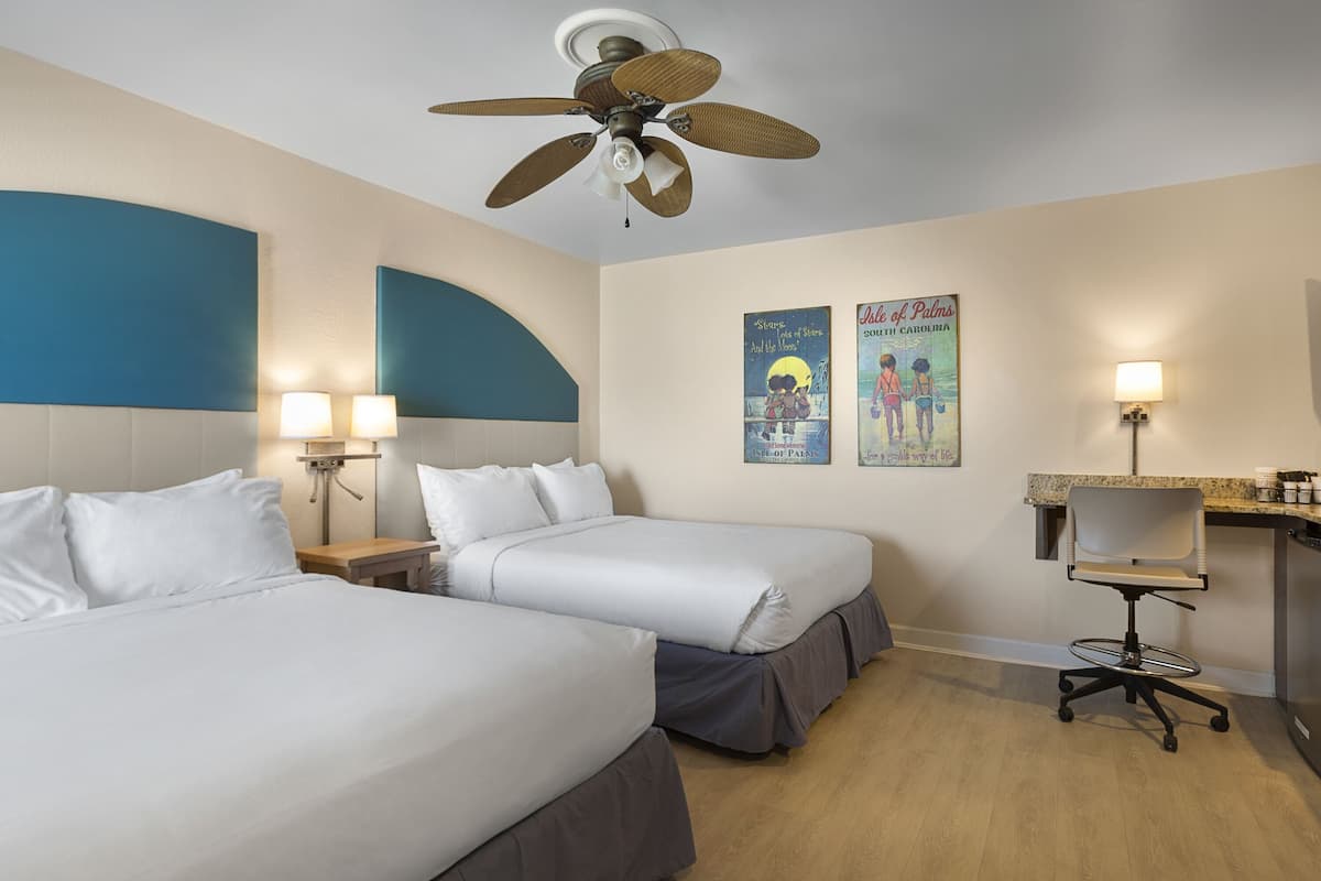 A hotel room with two beds, a desk, a chair, a ceiling fan, and wall art.