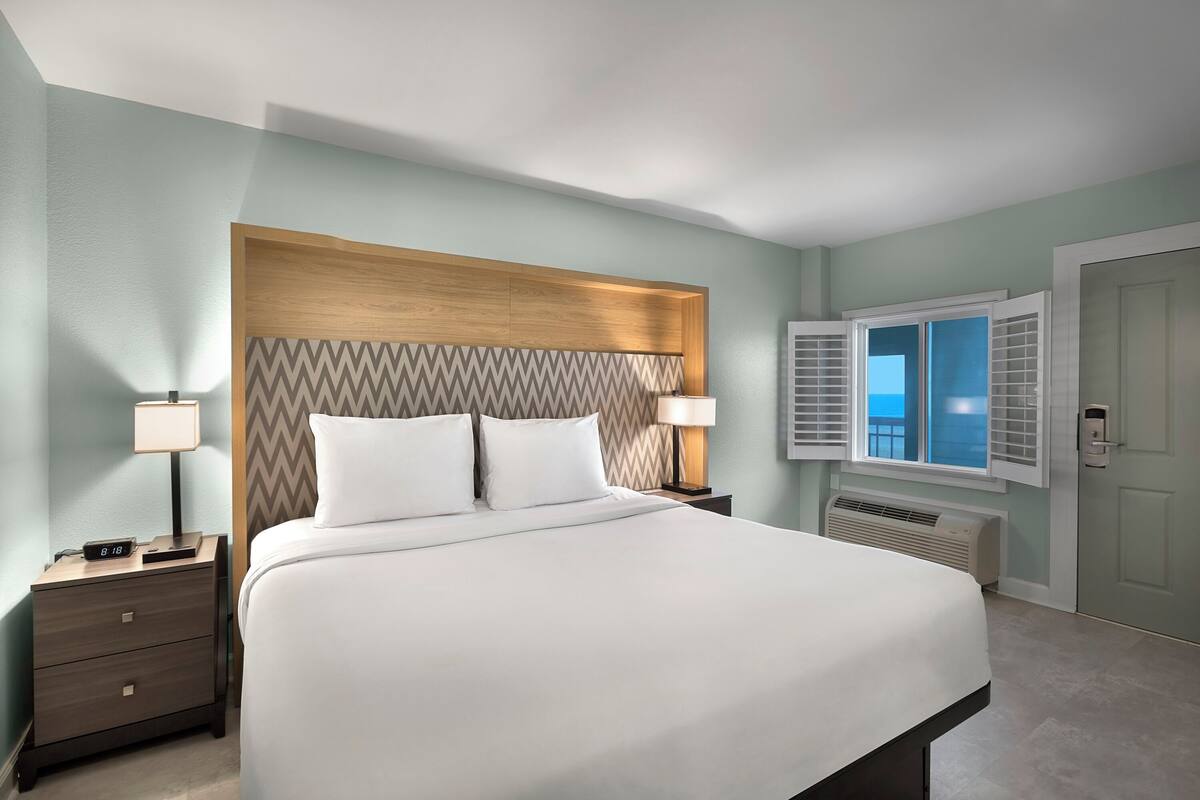 A neatly made bed with a wooden headboard, two bedside tables with lamps, a window with shutters, and a door.