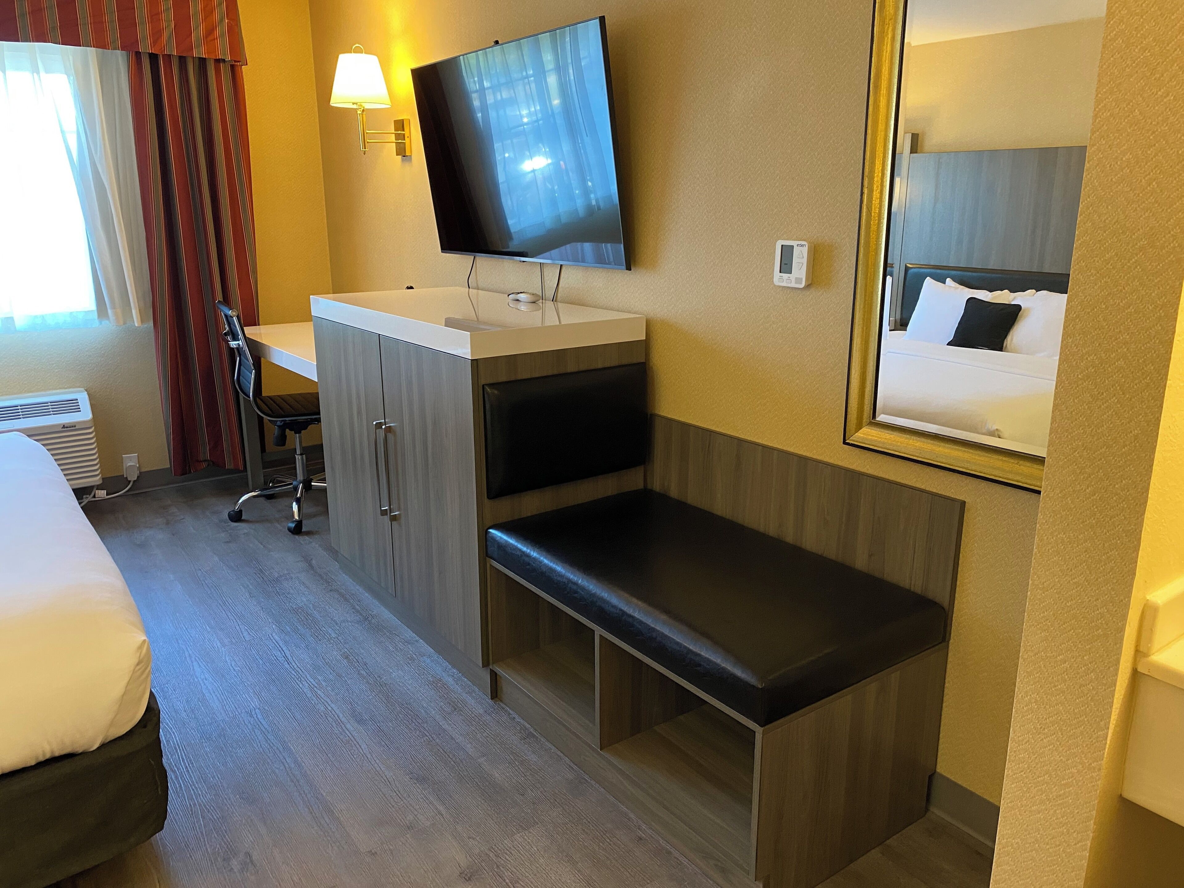 A hotel room with a bed, a desk, a TV, a bedside table, a chair, and a mirror.
