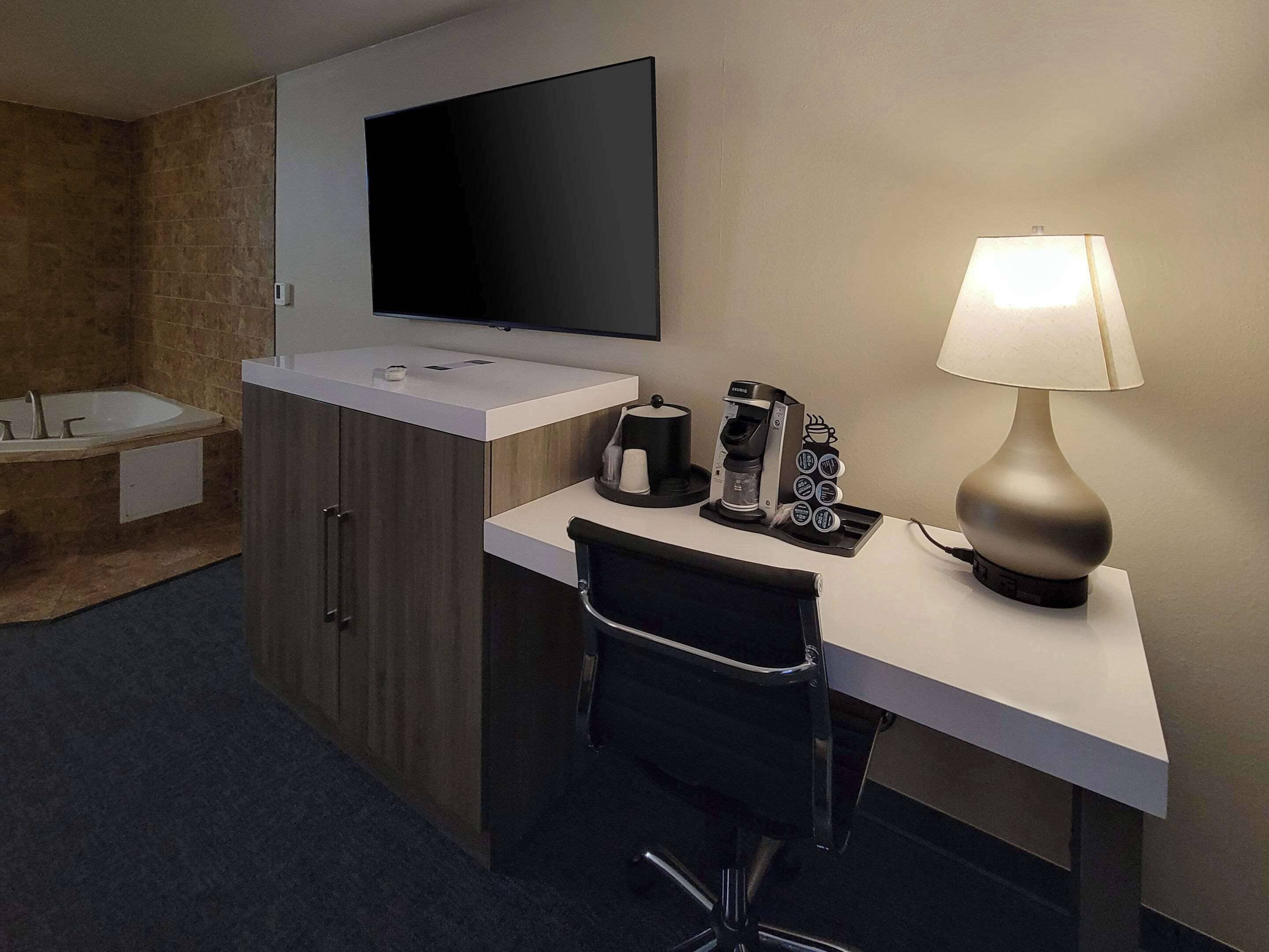 A hotel room with a flat-screen TV mounted on the wall, a desk with a lamp, a coffee maker, and a chair.