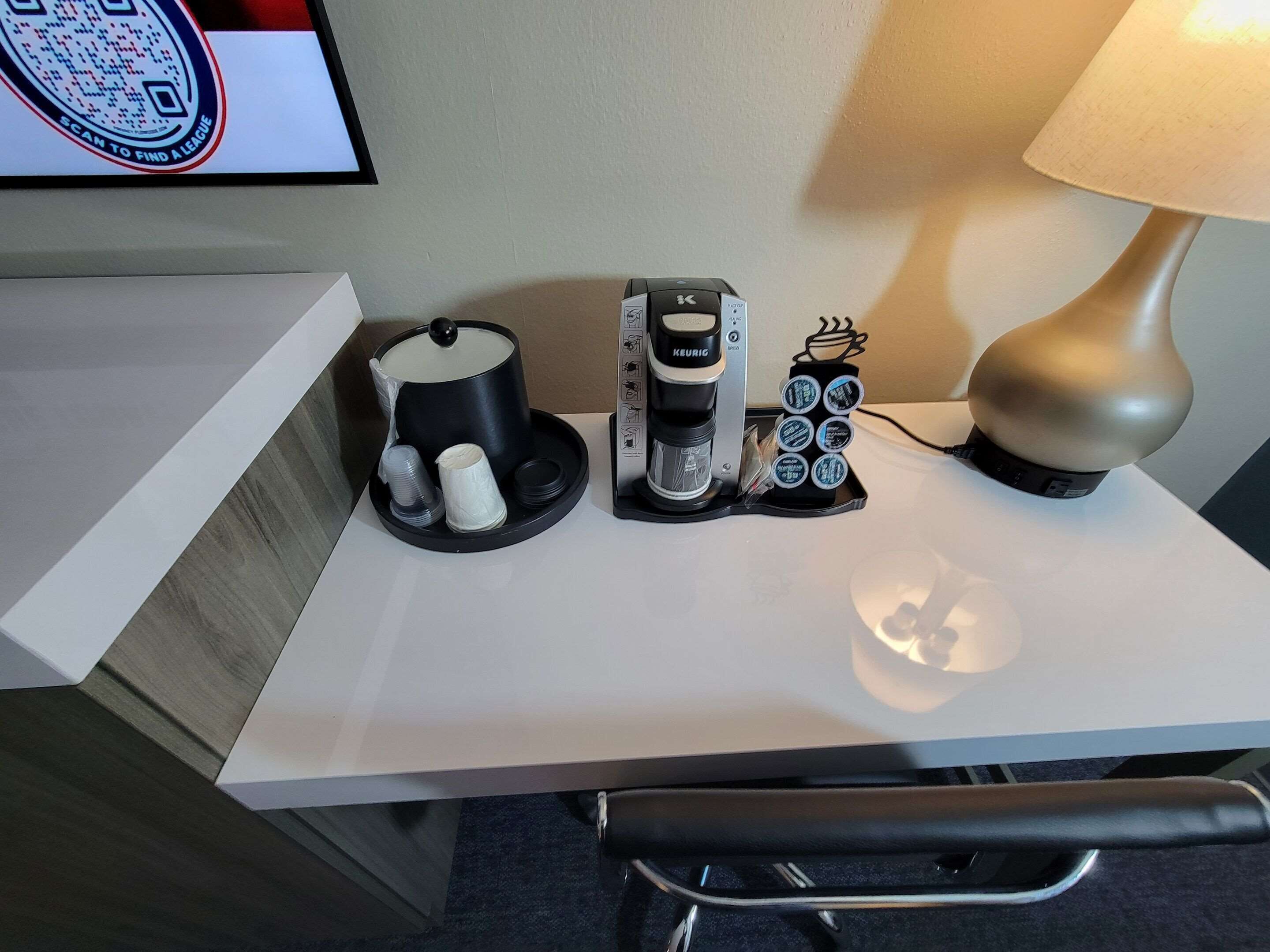 Dans une chambre d’hôtel, on trouve un bureau équipé d’une machine à café Keurig, d’une cafetière, d’un porte-tasse et d’une lampe.