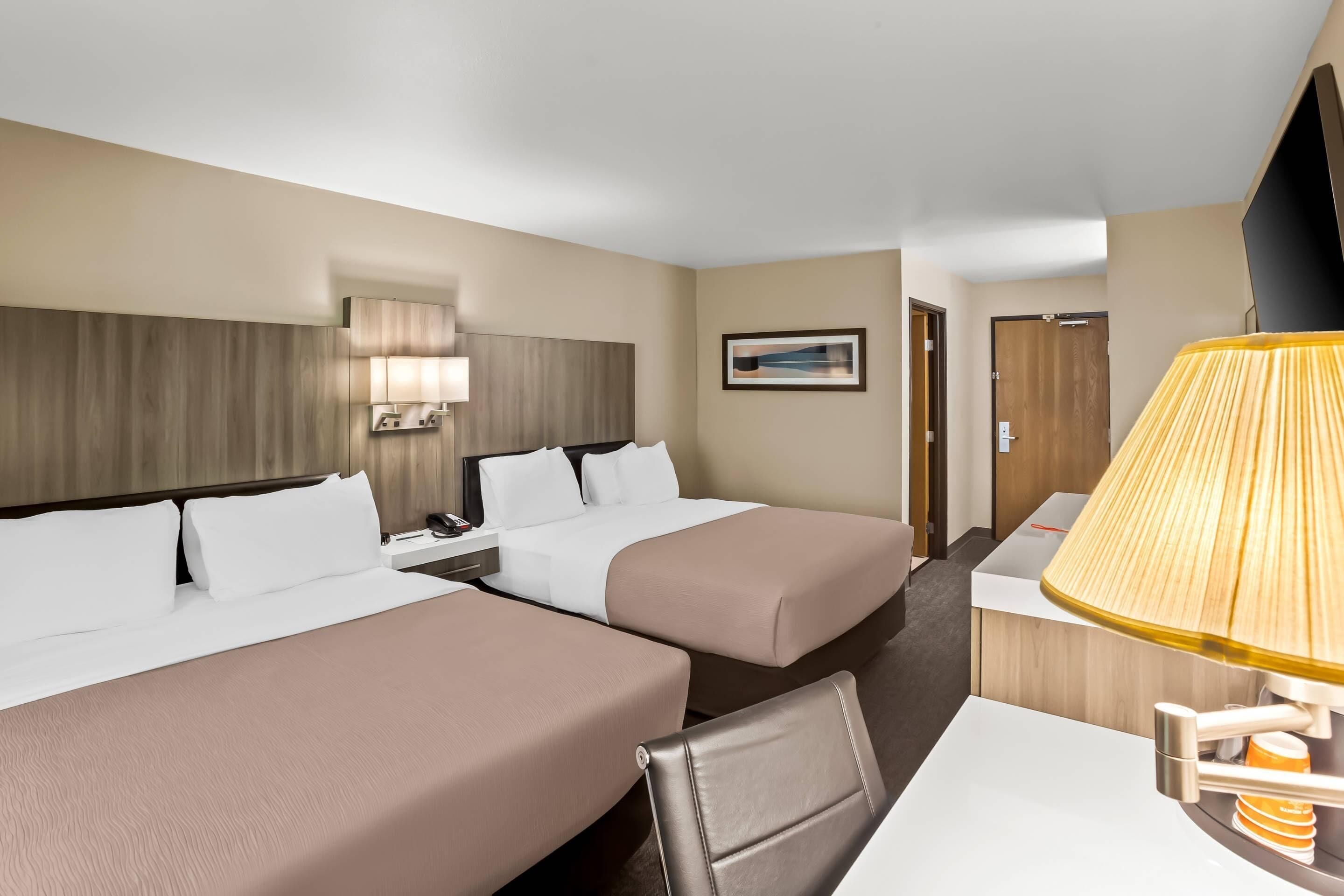A hotel room with two beds, a desk, a lamp, a TV, and a painting on the wall.