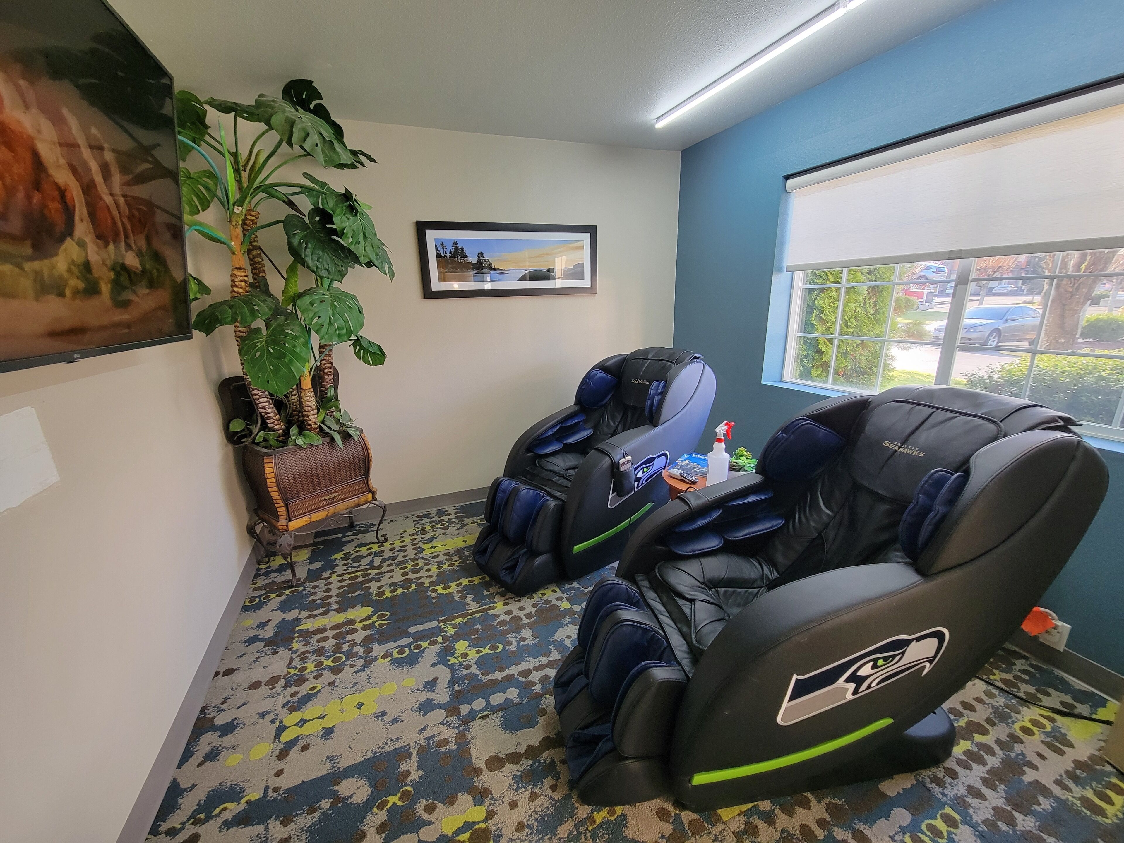 A room with two massage chairs, a large potted plant, a framed picture on the wall, and a window with a view of the outside.