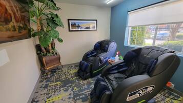 A room with two massage chairs, a large potted plant, a framed picture on the wall, and a window with a view of the outside.
