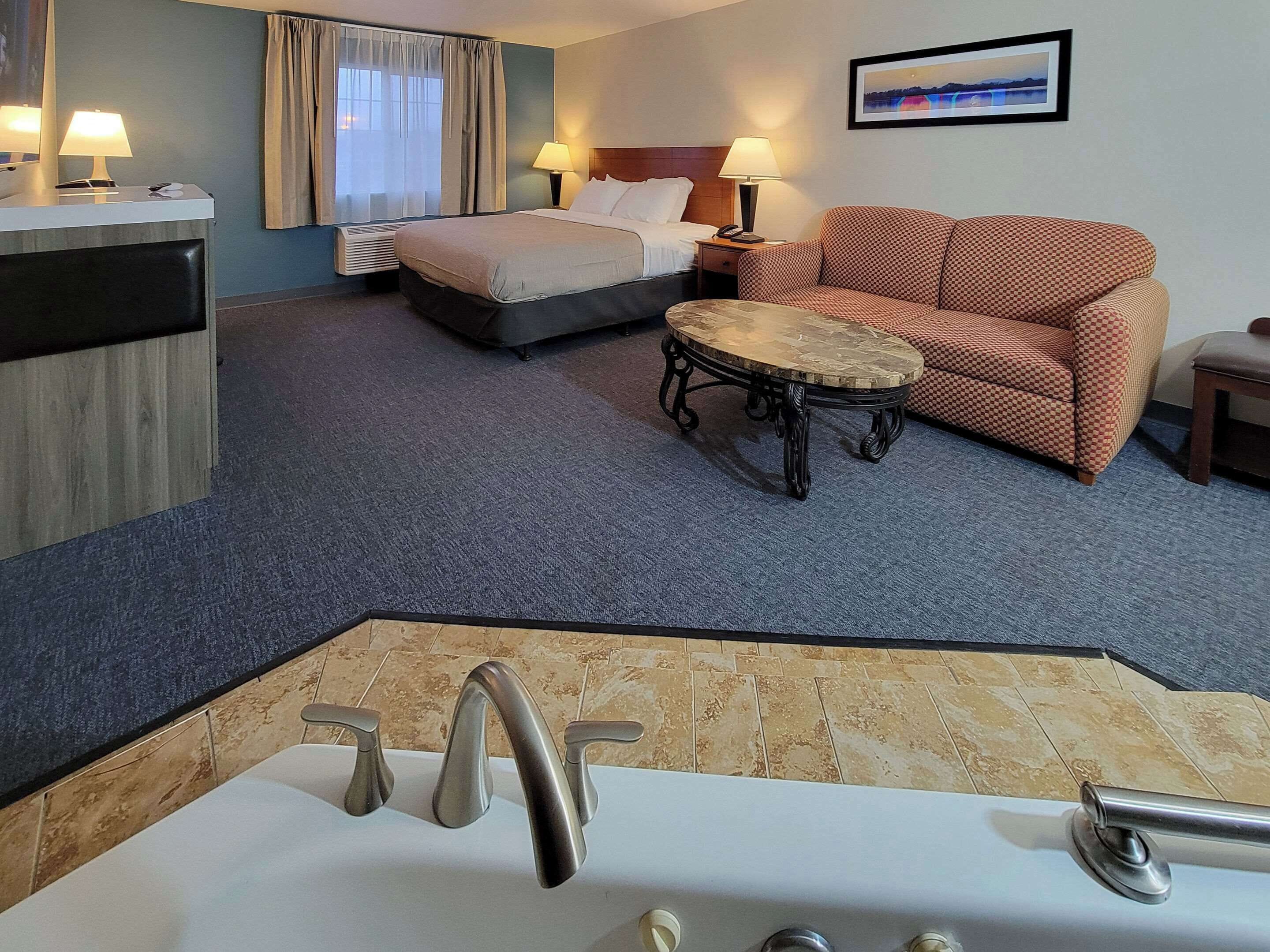 A hotel room with a bed, a sofa, a small round table, a desk with a lamp, a bathroom with a bathtub, and a framed picture on the wall.