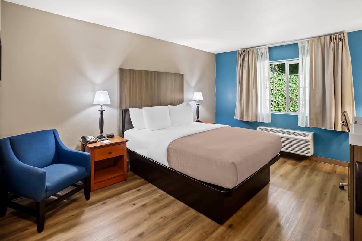 Une chambre d’hôtel avec un lit, des tables de chevet, une chaise, une fenêtre avec des rideaux et un radiateur.