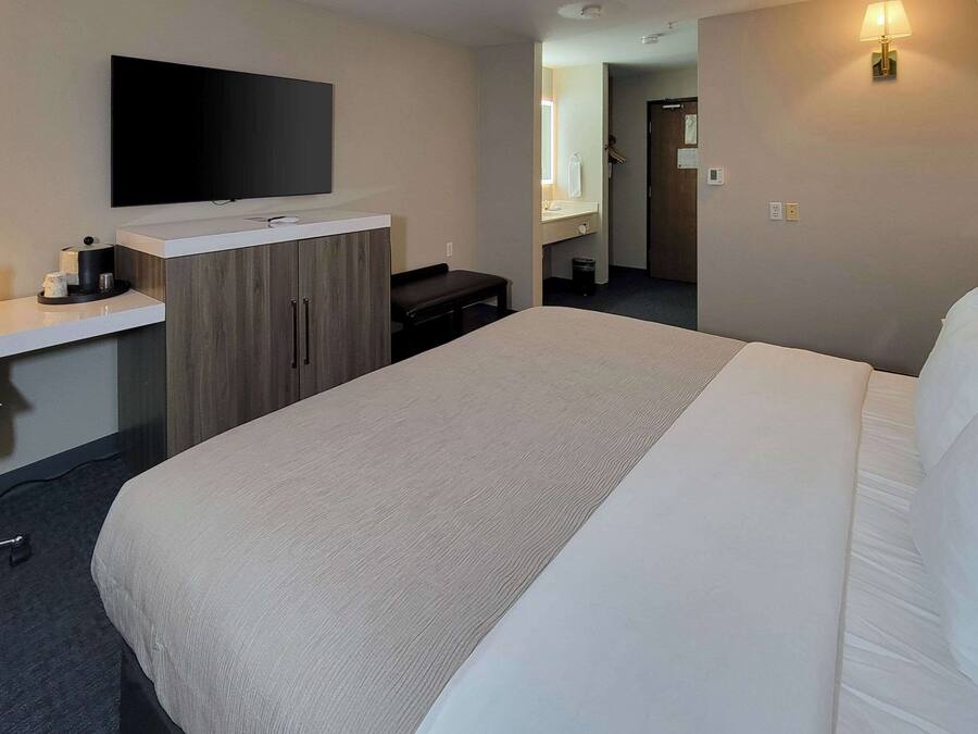 Una camera d'albergo con un letto, una TV a parete, una piccola scrivania con lampada e uno specchio.