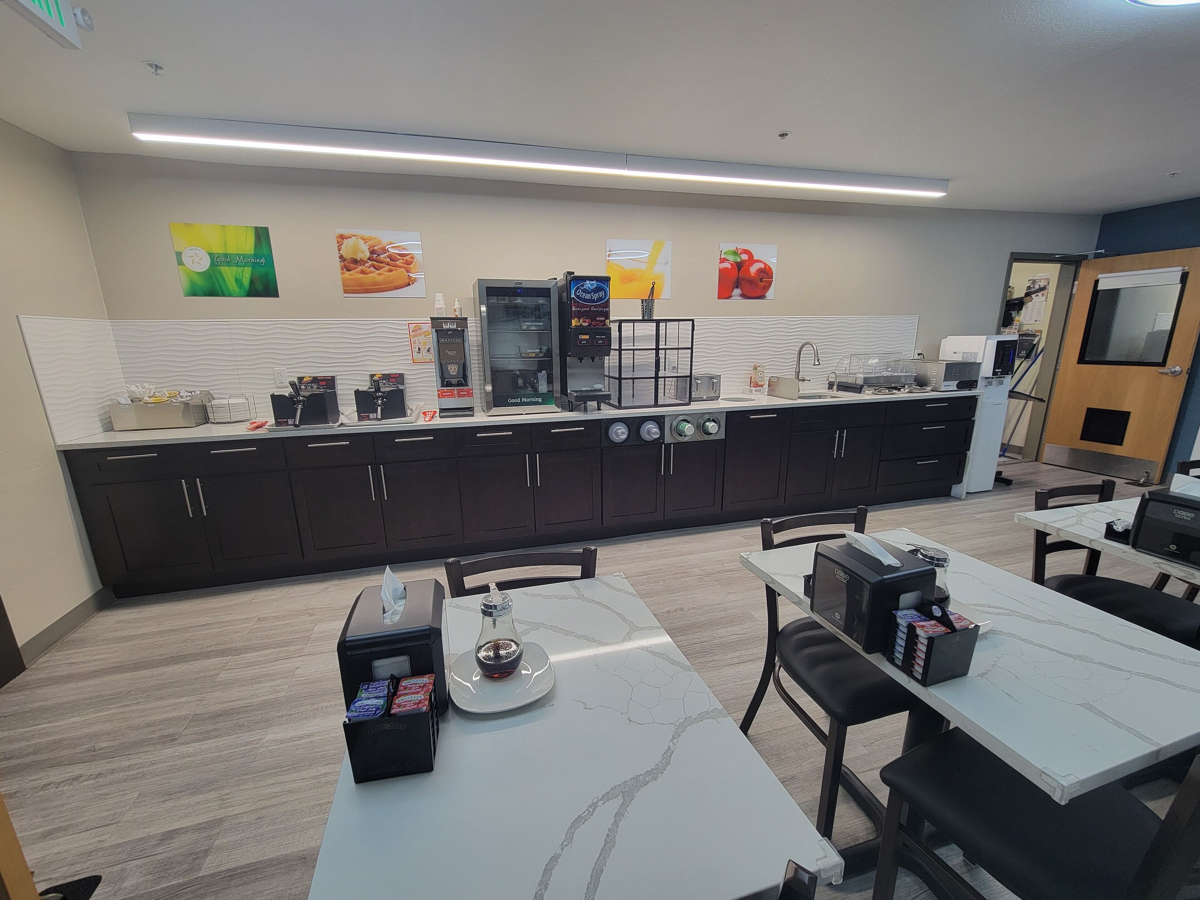Een moderne keuken met een lange aanrecht, voorzien van koffieautomaten, een magnetron en een koelkast. Er staan meerdere tafels en stoelen klaar voor een maaltijd.