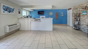 A reception area with a blue accent wall, a white reception desk, a mounted TV, and nautical decor including a ship's wheel and anchor. - Ocean's Edge Hotel, Port Aransas,TX (Port Aransas)