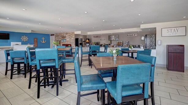 A modern dining area with blue upholstered chairs and tables, a kitchen in the background, and a sign that says "STAY and be nice." - Ocean's Edge Hotel, Port Aransas,TX (Port Aransas)