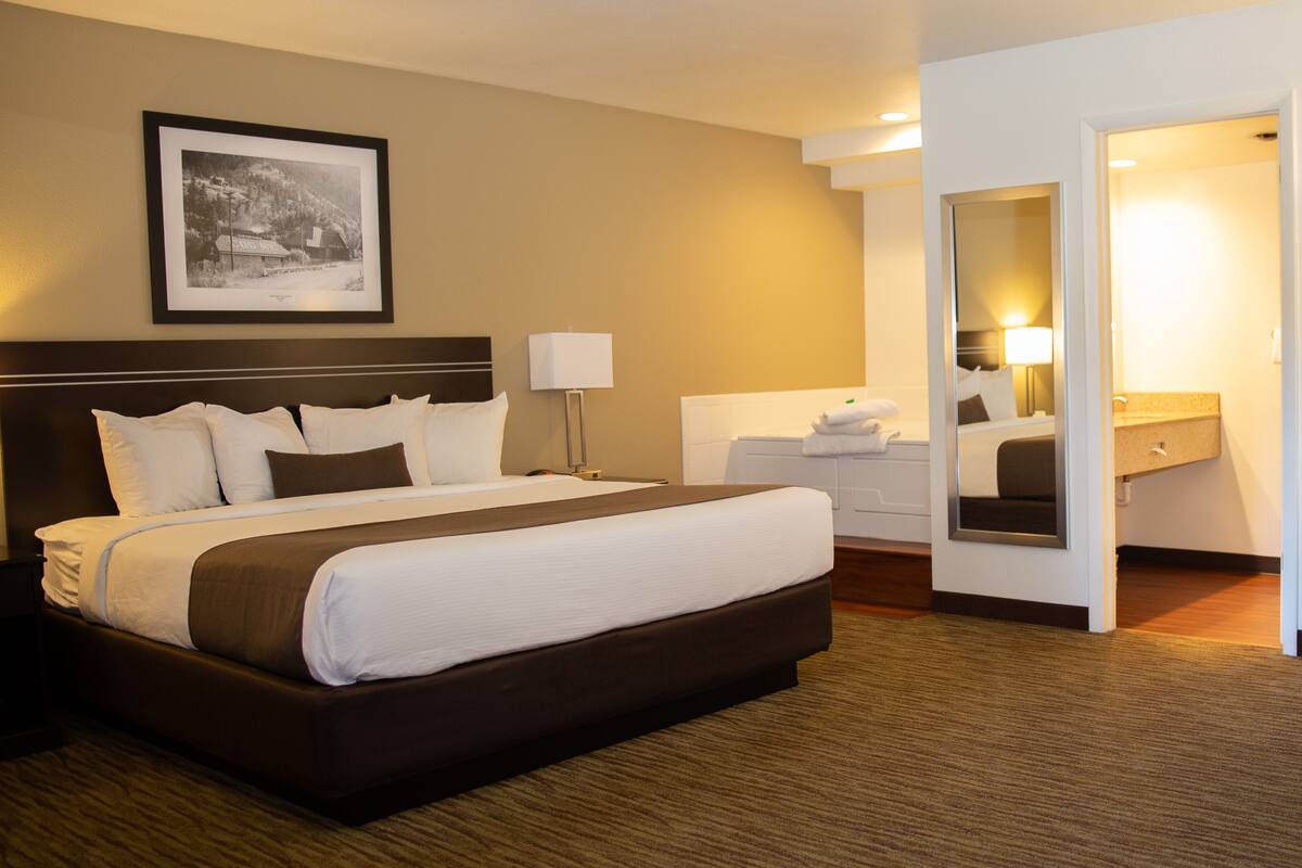 A hotel room with a large bed, a bedside table, a lamp, a framed picture on the wall, and a bathroom visible through an open door.