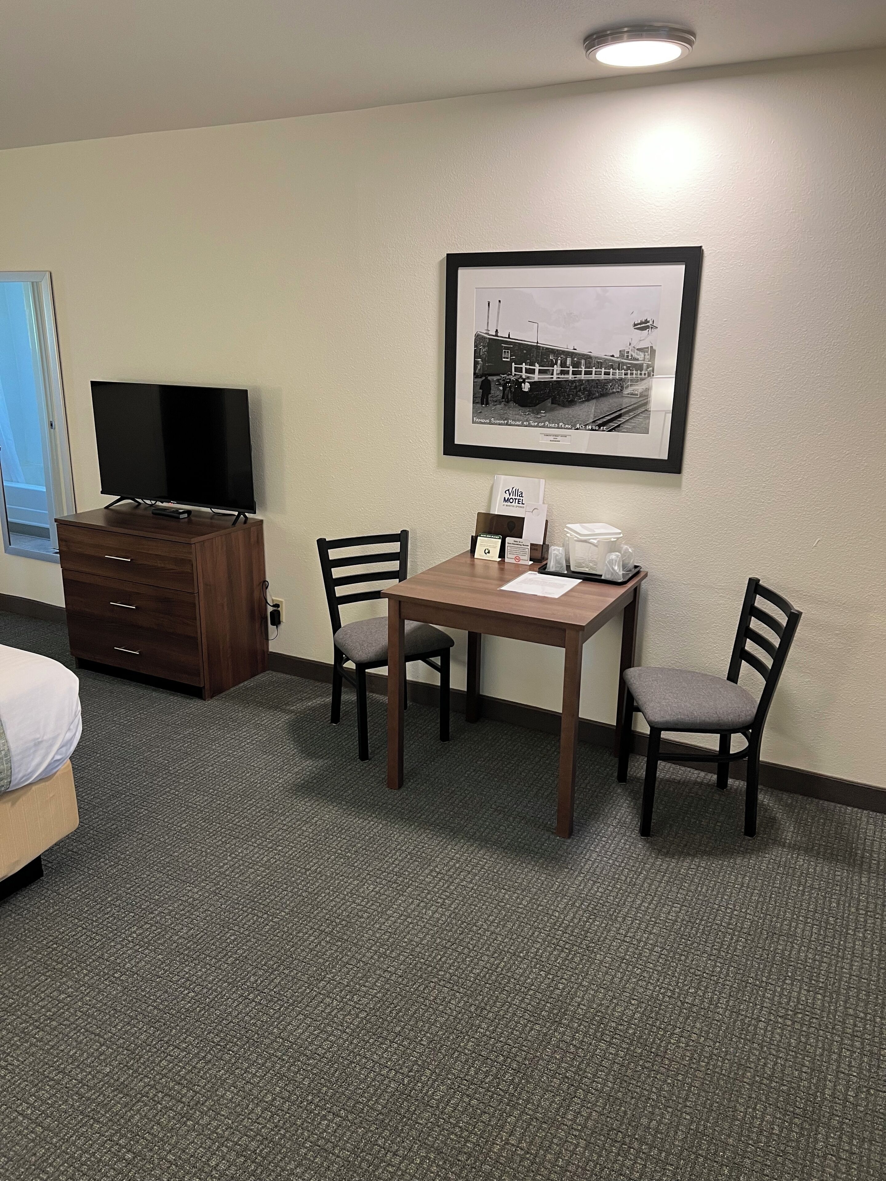 Una habitación de hotel con una cama, un televisor en una cómoda, una mesa de comedor con sillas, un cuadro en la pared y un minibar.