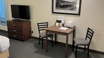 Una habitación de hotel con una cama, un televisor en una cómoda, una mesa de comedor con sillas, un cuadro en la pared y un minibar.