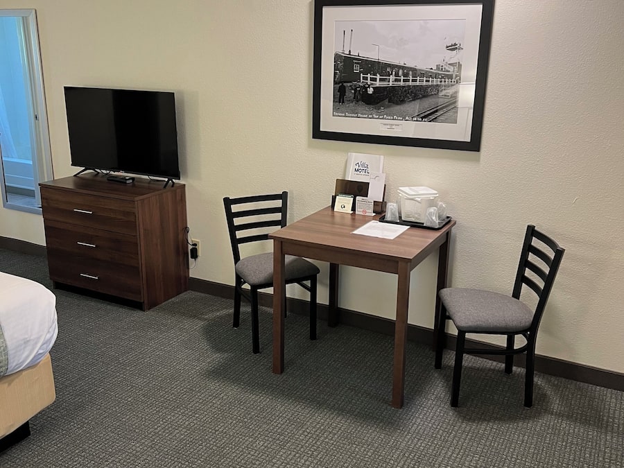 Una habitación de hotel con una cama, un televisor en una cómoda, una mesa de comedor con sillas, un cuadro en la pared y un minibar.