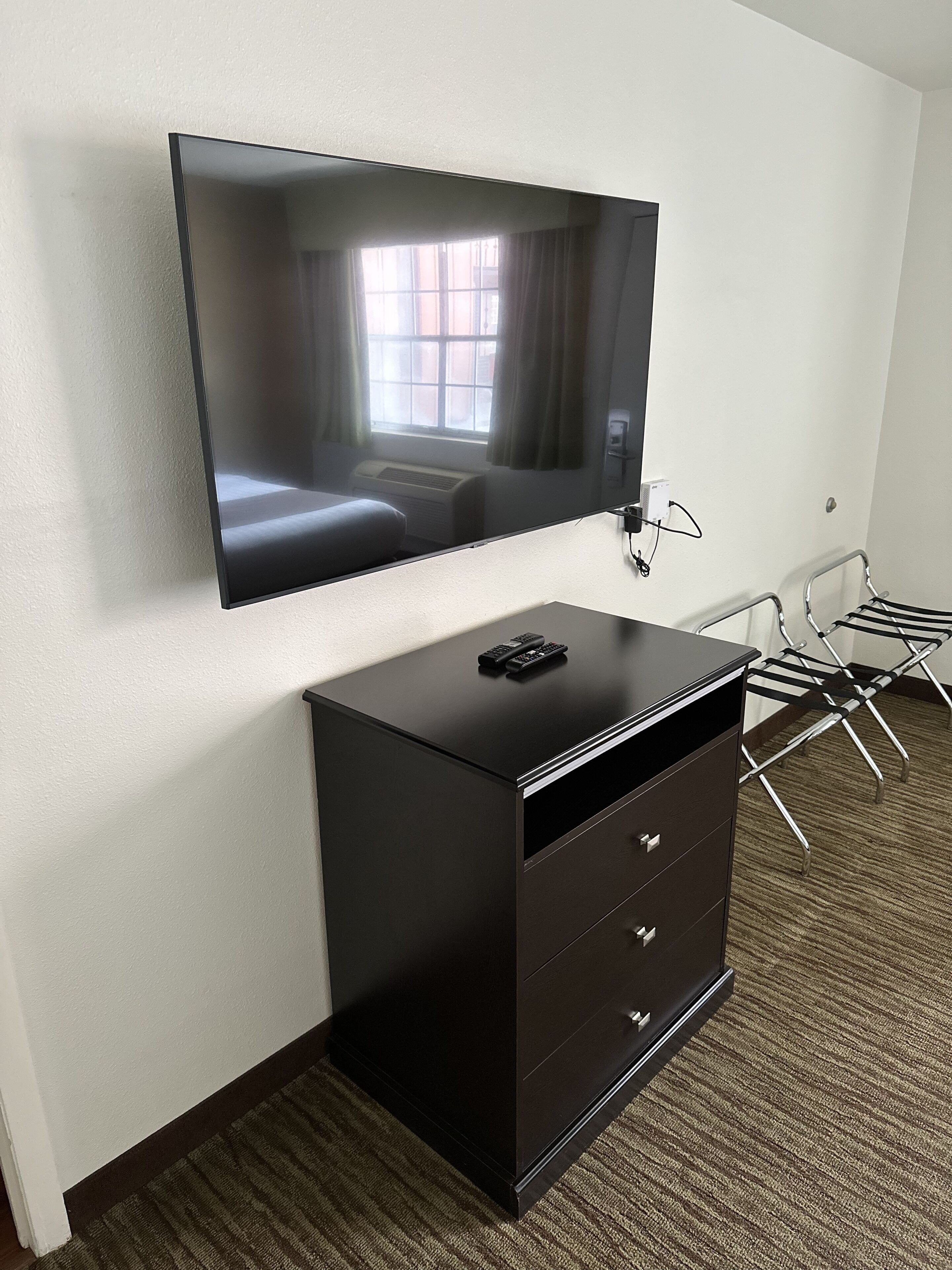 A hotel room with a flat-screen TV mounted on the wall, a black dresser with drawers, and a metal chair with a towel rack.