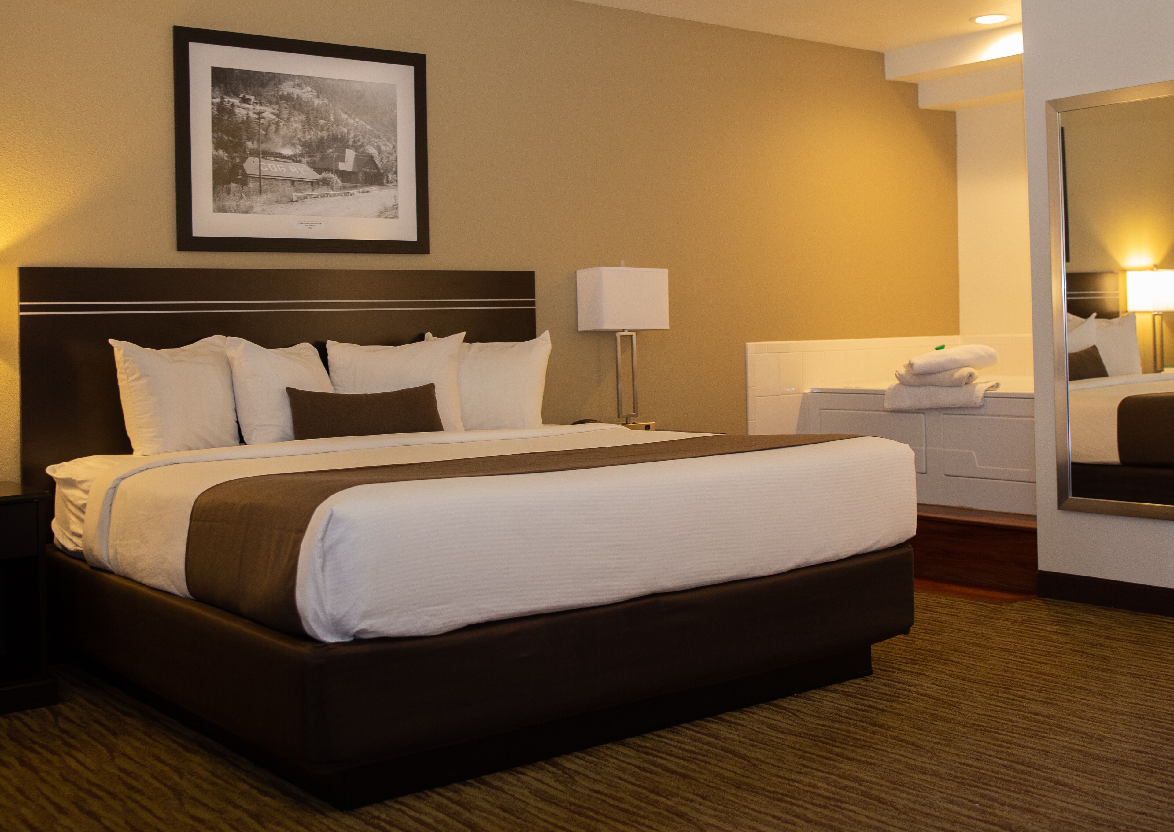 A hotel room with a large bed, a bedside table, a lamp, a framed picture on the wall, and a bathroom with a mirror and towel rack.