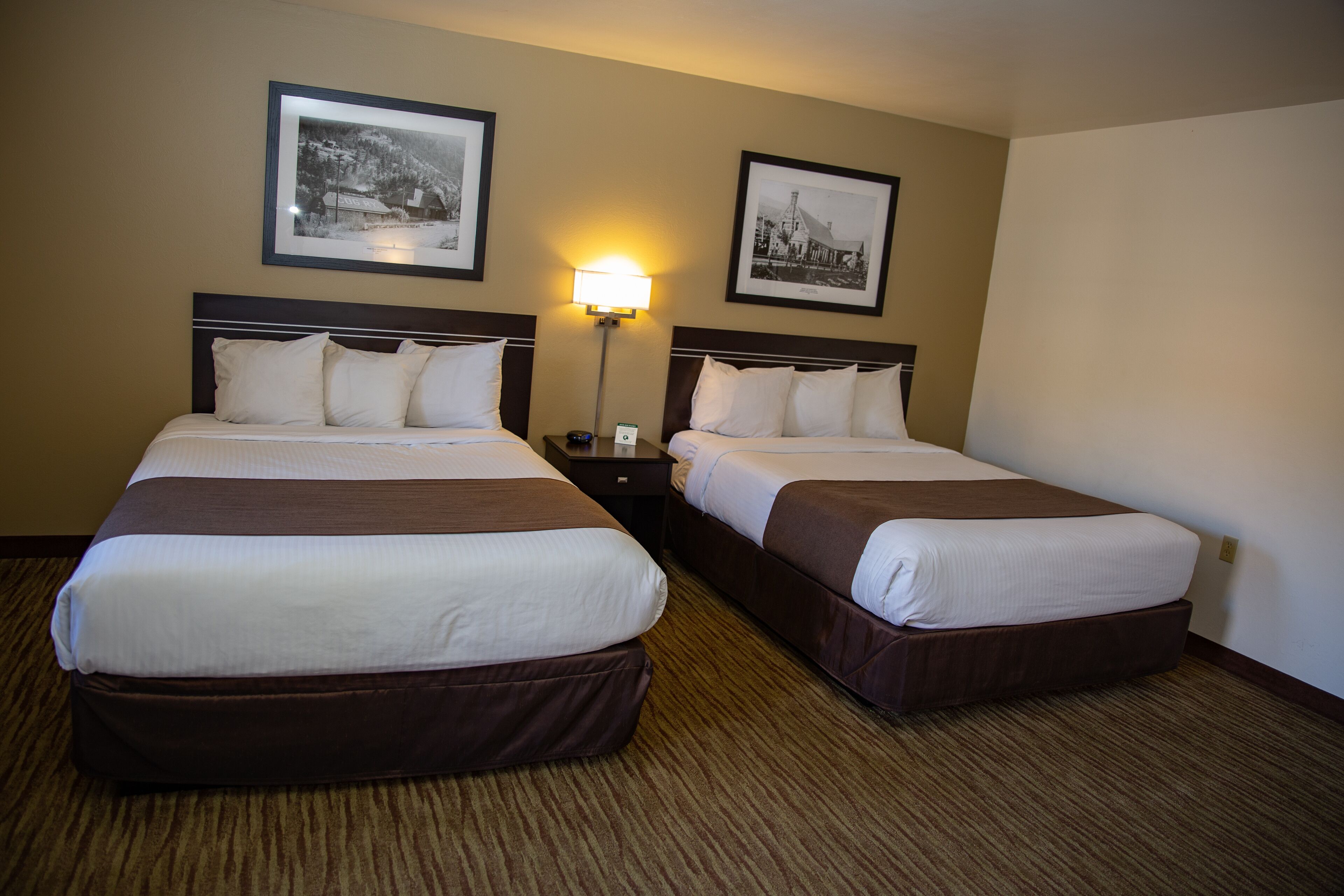 A hotel room with two beds, a nightstand, a lamp, and two framed pictures on the wall.