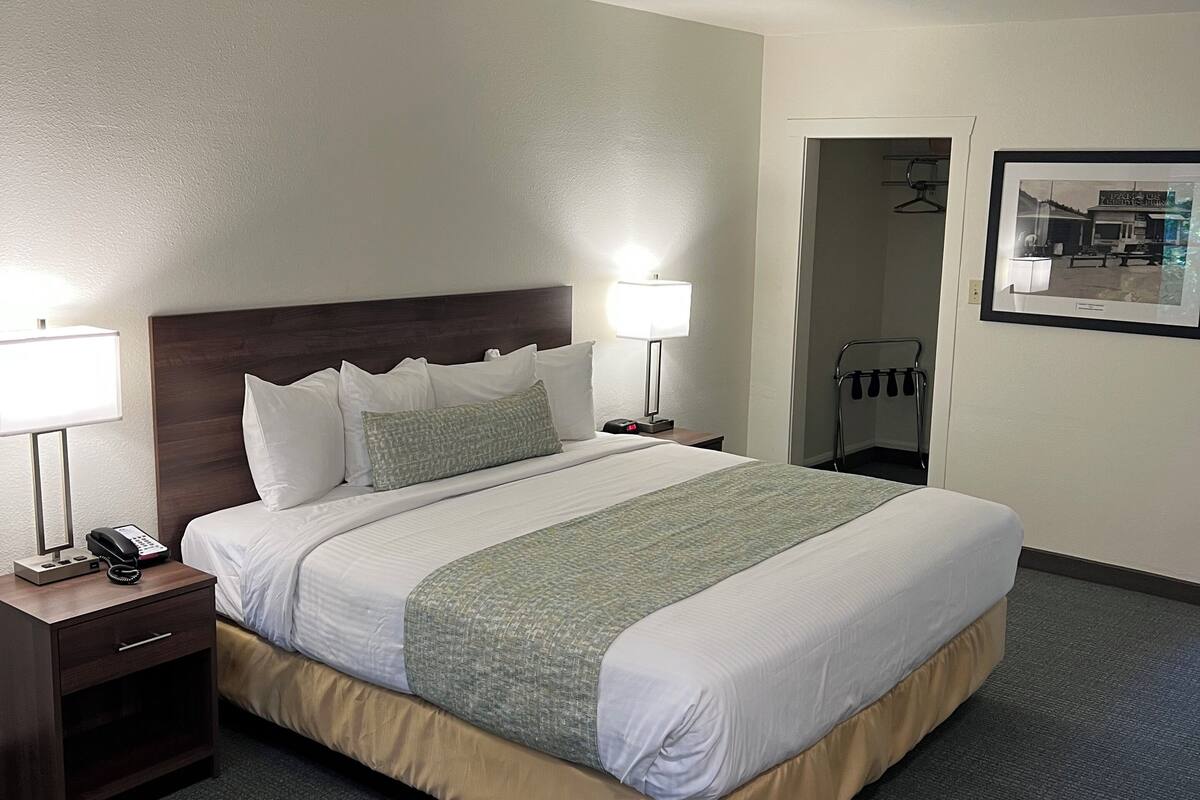 A hotel room with a large bed, two bedside tables with lamps, a nightstand, a framed picture on the wall, and a door leading to a bathroom.