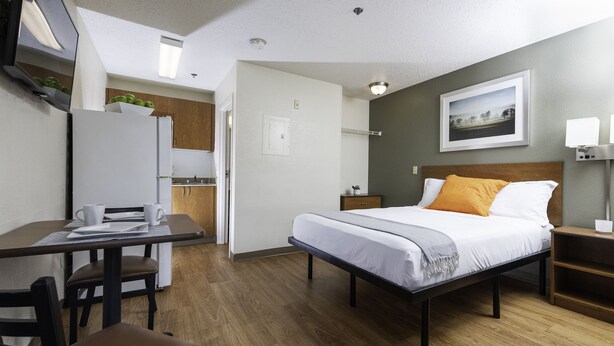 A compact hotel room with a bed, bedside table, and a desk with chairs. - Siegel Select Baton Rouge (Baton Rouge)