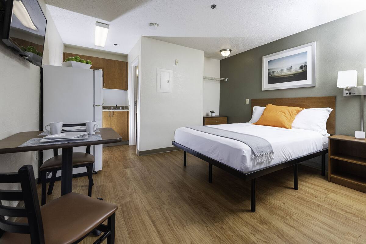 A compact hotel room with a bed, bedside table, and a desk with chairs.