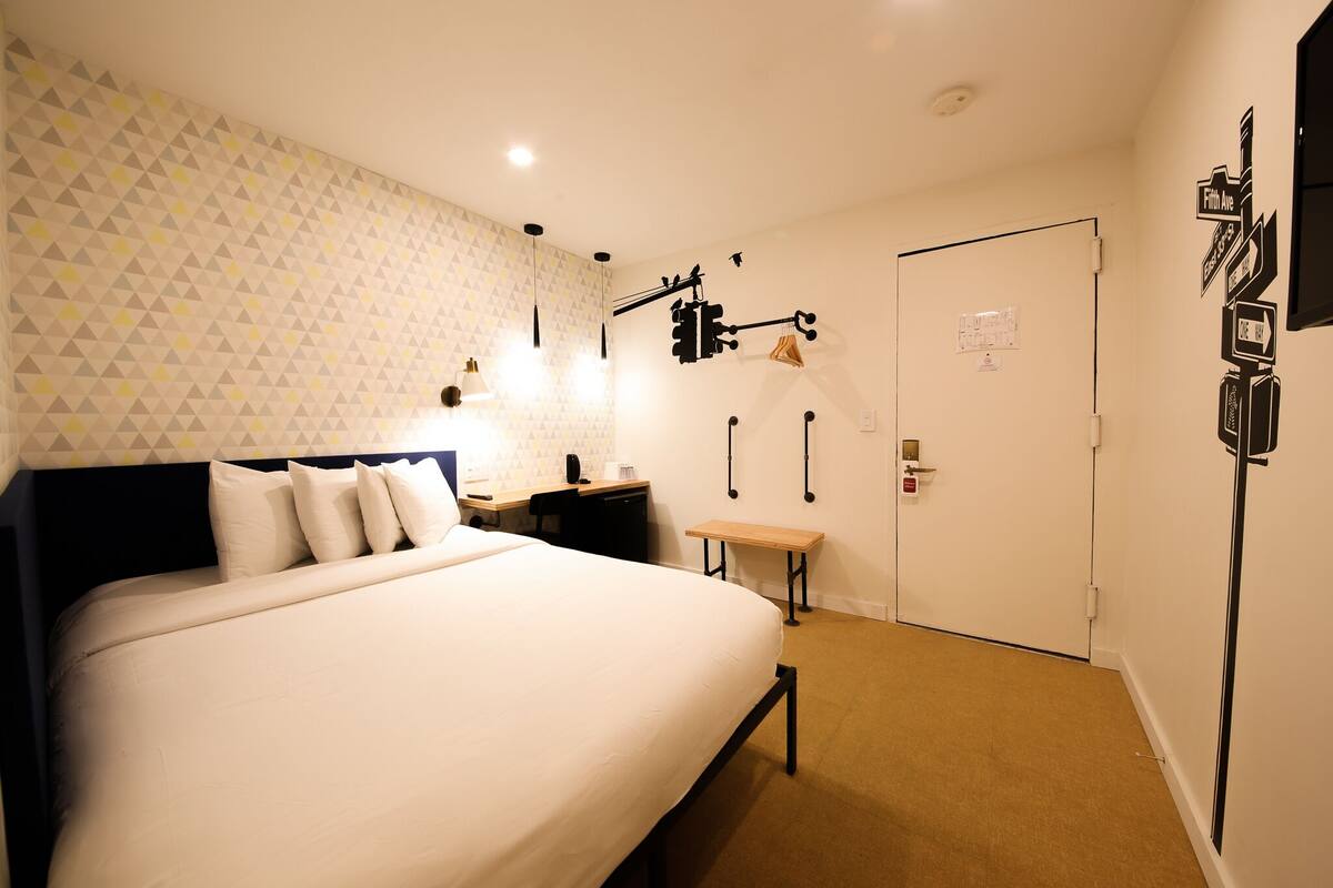 A hotel room with a bed, bedside table, lamp, and wall-mounted hooks.