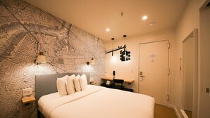 A hotel room with a large map on the wall, a bed with white linens, a desk with a lamp, and a door. - Doxie Hotel (New York)