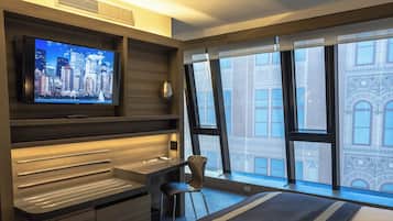Una camera d'albergo moderna con un grande televisore su una mensola in legno, una scrivania con una sedia e un'ampia finestra con vista sulla città.