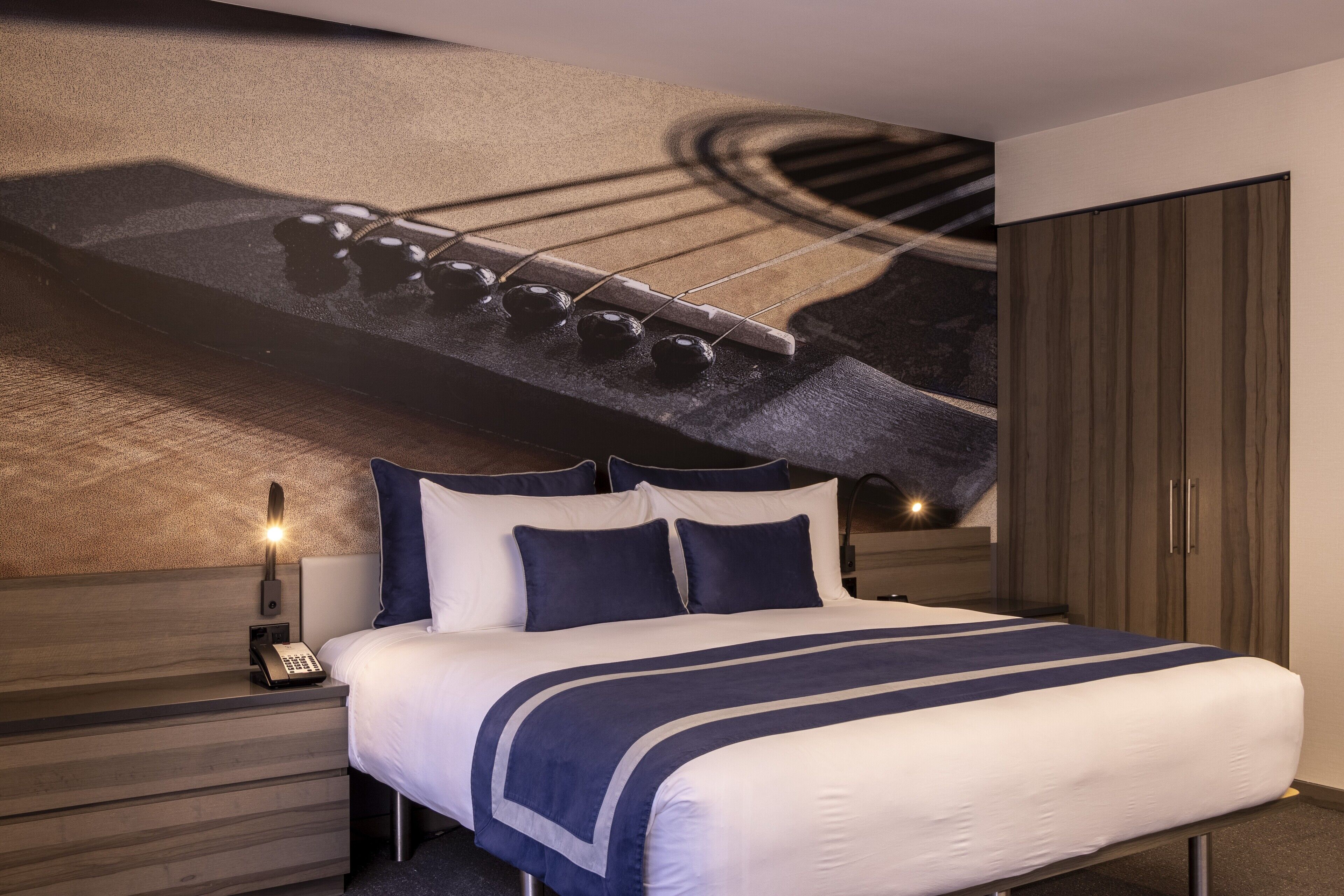 Une chambre d’hôtel comprenant un lit, une table de chevet avec un téléphone, une lampe fixée au mur et une armoire.