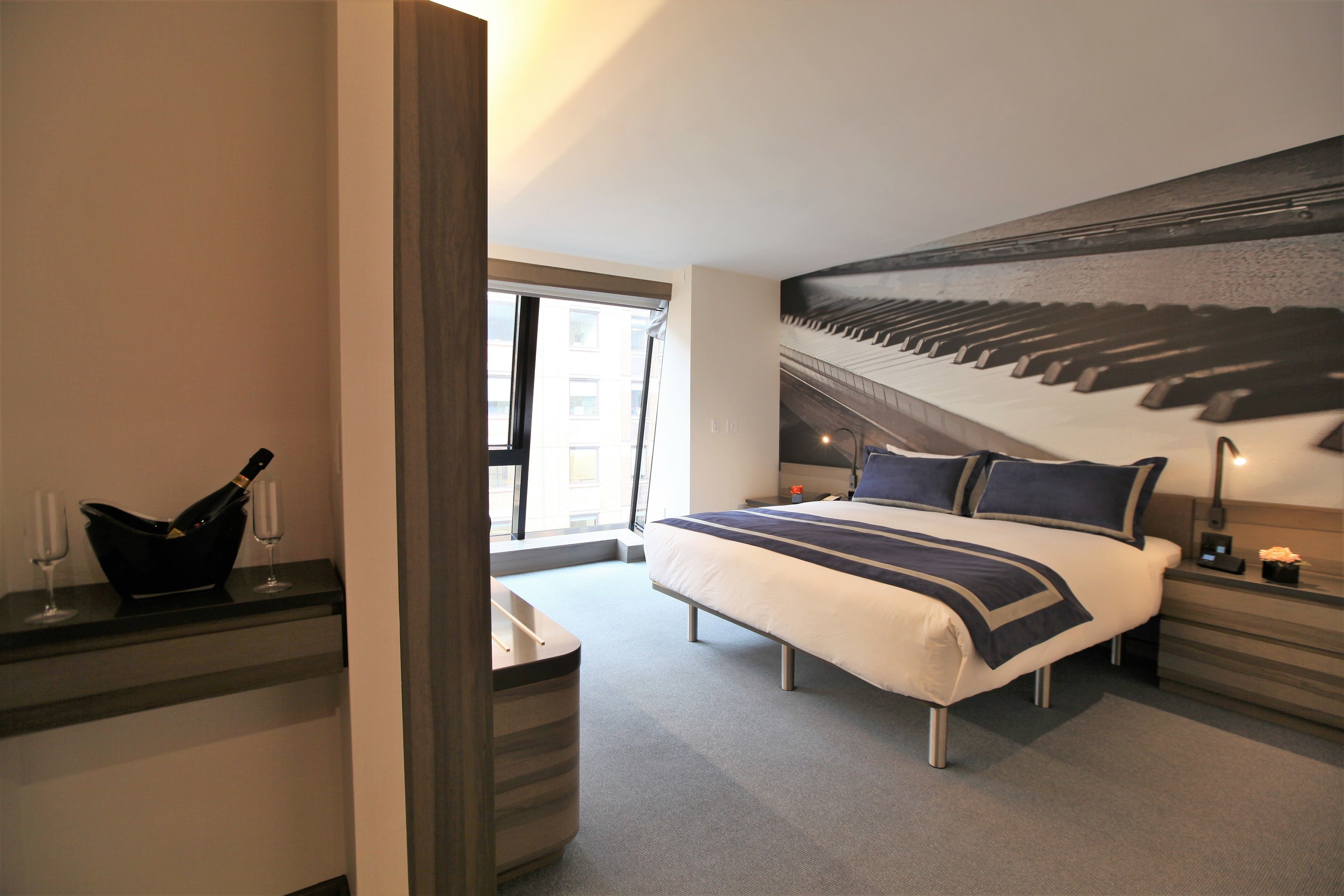 Quarto de hotel moderno com uma cama grande, arte de parede com um piano, uma pequena mesa com uma garrafa e copos, e vista da cidade pela janela.