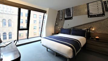 A modern hotel room with a large bed, a desk with a laptop, and a view of the cityscape through large windows.