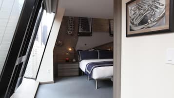 Quarto compacto com uma cama, uma secretária pequena e uma janela com vista para a cidade.