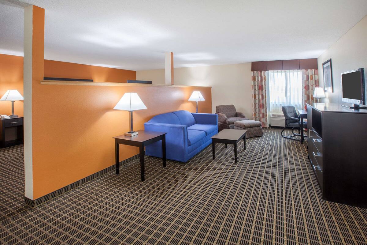 A hotel room with a blue sofa, two lamps, a desk, a TV, and a patterned carpet.
