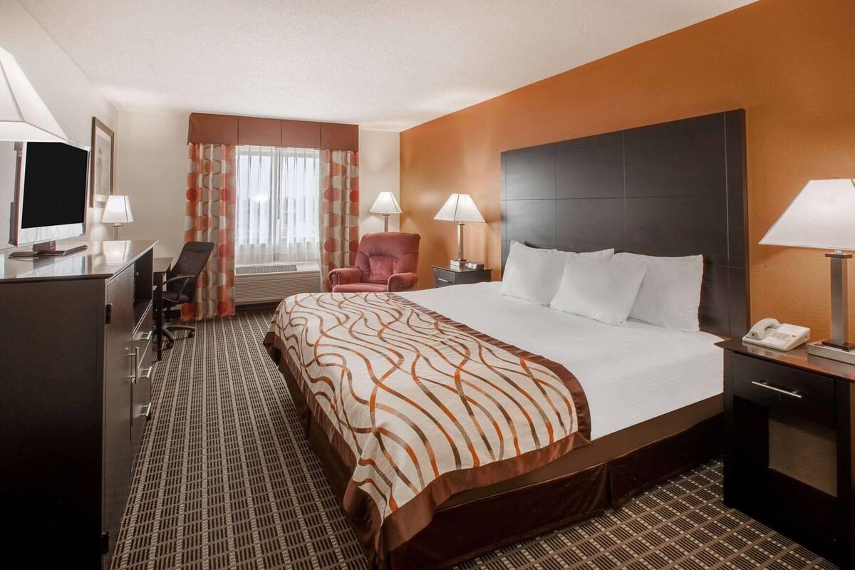 A hotel room with a large bed, a desk with a computer, a chair, a desk lamp, a telephone, and a window with curtains.