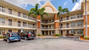 A multi-story hotel with a central atrium, palm trees, and a parking lot with two cars. - Extended Stay America Suites Ft Lauderdale Cyp Crk Andrews A (Oakland Park)