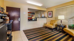 A hotel lobby with a reception desk, a coffee machine, a sofa, a small table with a lamp, and a striped carpet. - Extended Stay America Suites Ft Lauderdale Cyp Crk Andrews A (Oakland Park)