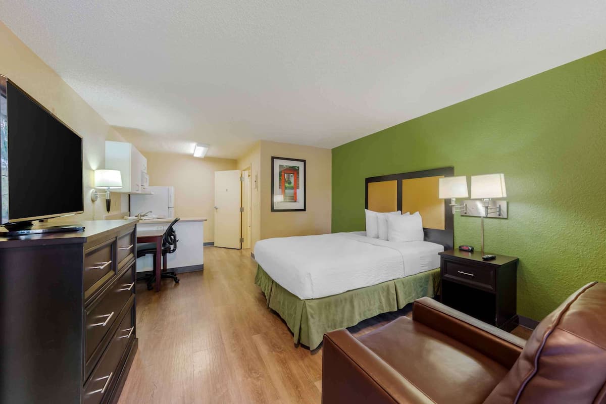 A hotel room with a bed, a desk with a computer, a chair, a TV, and a nightstand with a lamp.