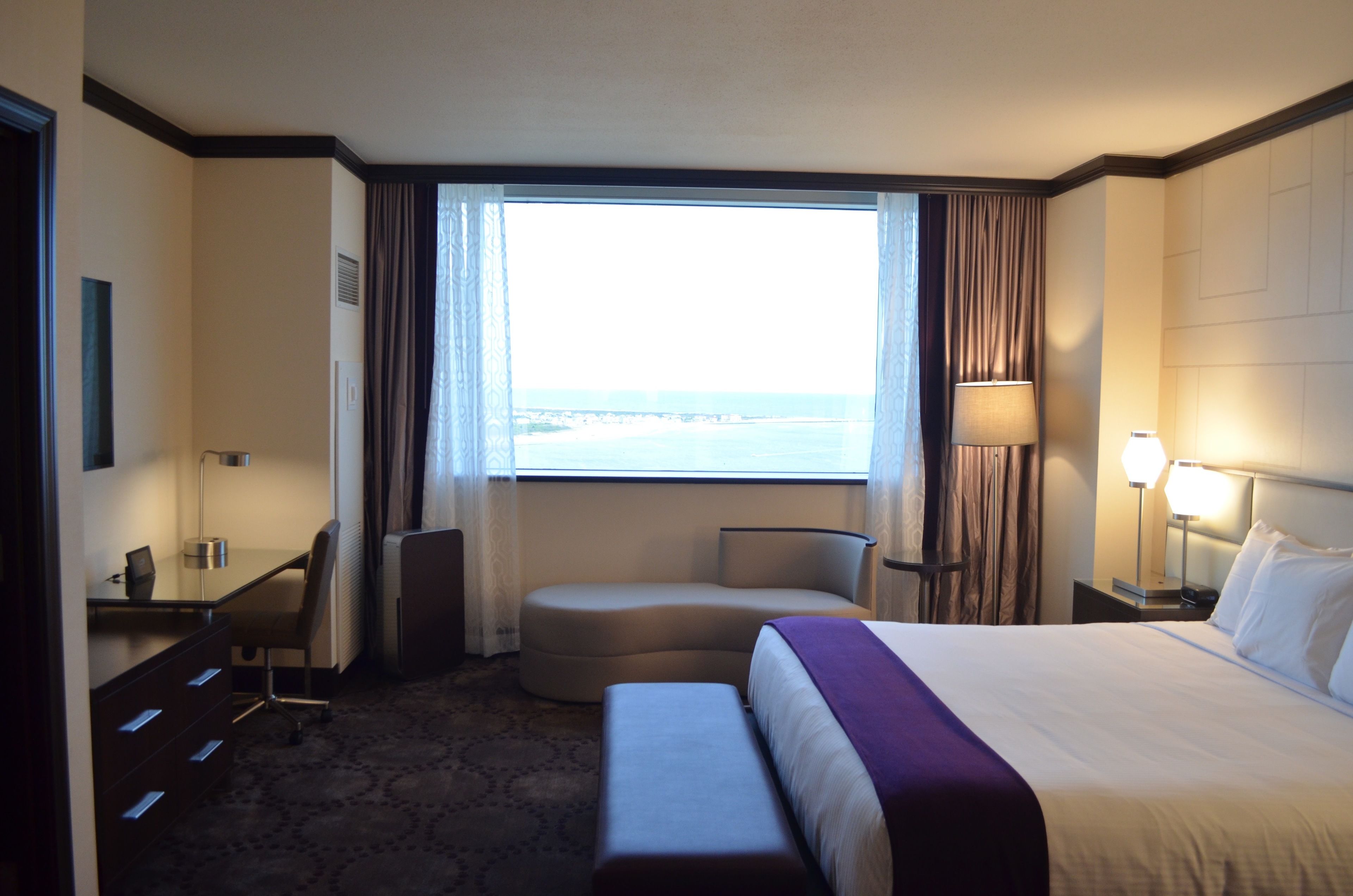 A hotel room with a bed, a desk, a chair, a small sofa, a lamp, and a view of the ocean.