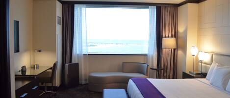 A hotel room with a bed, a desk, a chair, a small sofa, a lamp, and a view of the ocean.